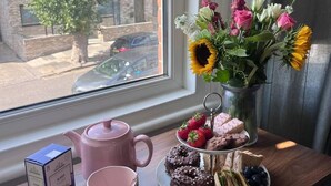 A table with a teapot, mugs, a plate of food, and a vase of flowers by a window. - Zozy Gladstone (London)