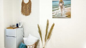 A room with a white wall, a painting of a woman at the beach, a woven wall hanging, a white refrigerator, a wicker basket, and pampas grass in a vase. - Zozy Gladstone (London)