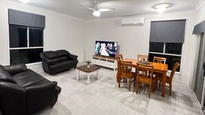 Een woonkamer met een zwarte leren bank, een houten eettafel met stoelen, een televisie en een plafondventilator.