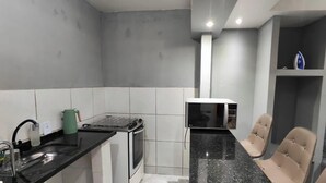 A compact kitchen with a stove, microwave, and sink. There are two chairs and a small shelf with a kettle. - Complete Studio 2 (Rio das Ostras)