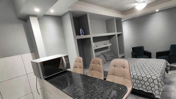 A modern bedroom with a bed, two chairs, a desk with a microwave, and built-in shelves. - Complete Studio 2 (Rio das Ostras)