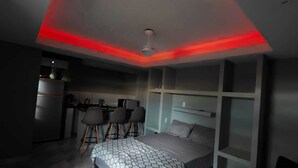 A modern bedroom with a bed, a desk, and chairs, and a kitchenette area. - Complete Studio 2 (Rio das Ostras)
