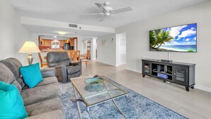 A living room with a sofa, coffee table, and TV. There is a ceiling fan, a kitchen in the background, and a dining area. - ☀Stunning Beachfront Condo🐬Price Drop!🌴Private Balcony (Indian Rocks Beach)