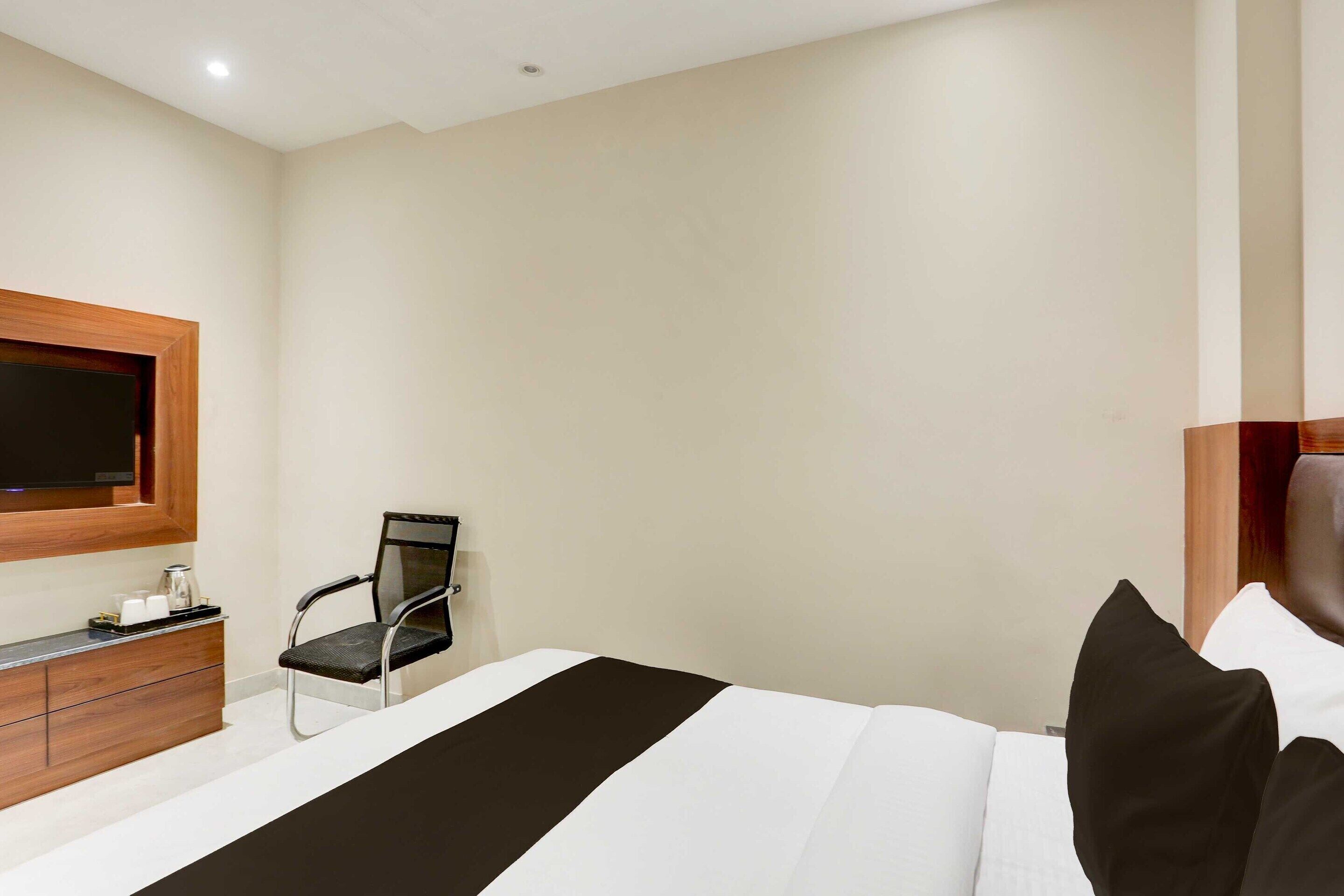 A hotel room with a bed, a TV mounted on the wall, a desk with a chair, and a small table with cups and a bottle.
