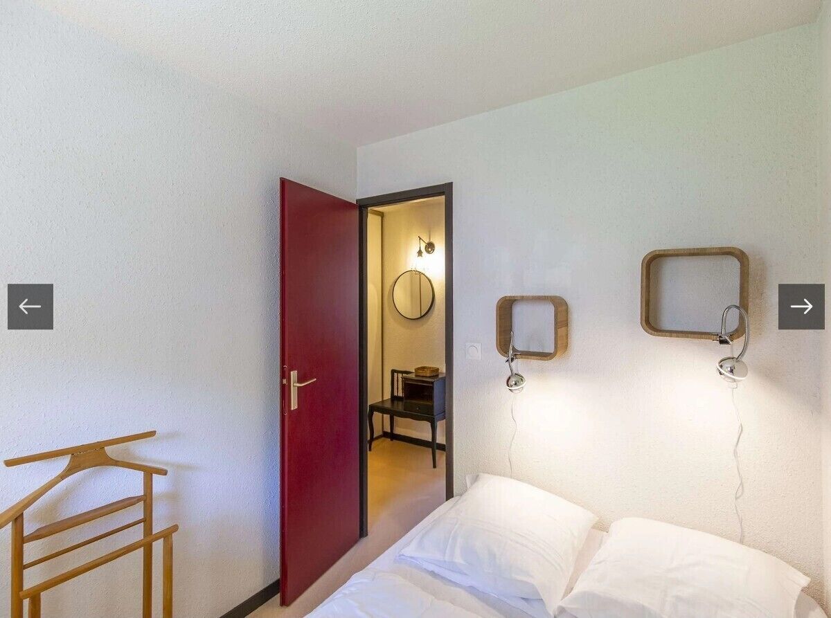 Una habitación de hotel con una cama, una silla de madera, una puerta roja y un espejo en la pared.