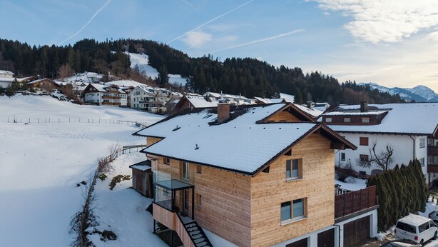 A two-story wooden house with a balcony, a staircase, and a garage with two cars parked inside. - New & high-quality furnished guest house in a quiet location - Burgberg im Allgäu (Burgberg im Allgäu)