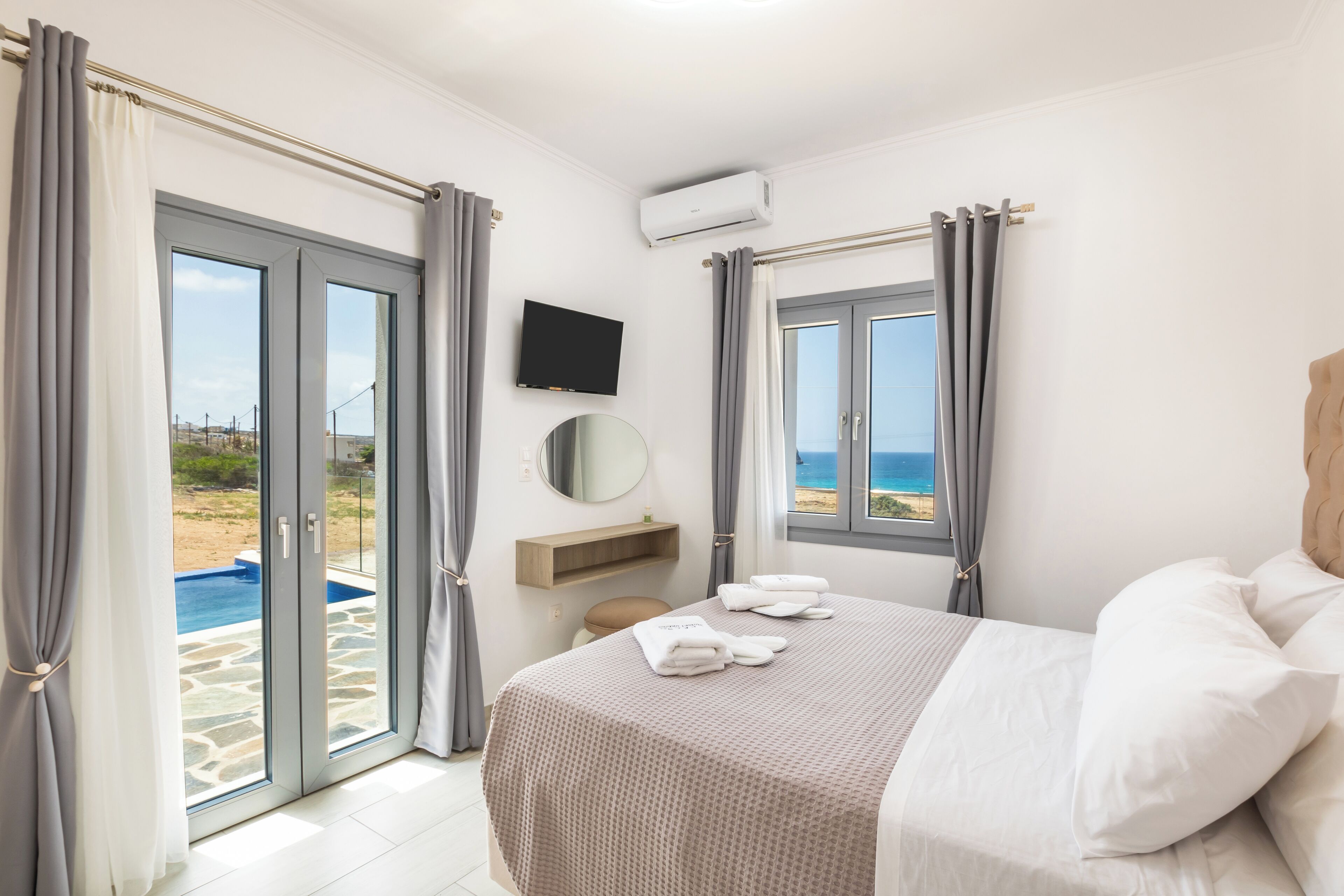 A bedroom with a bed, a TV, a view of the beach, and a pool.