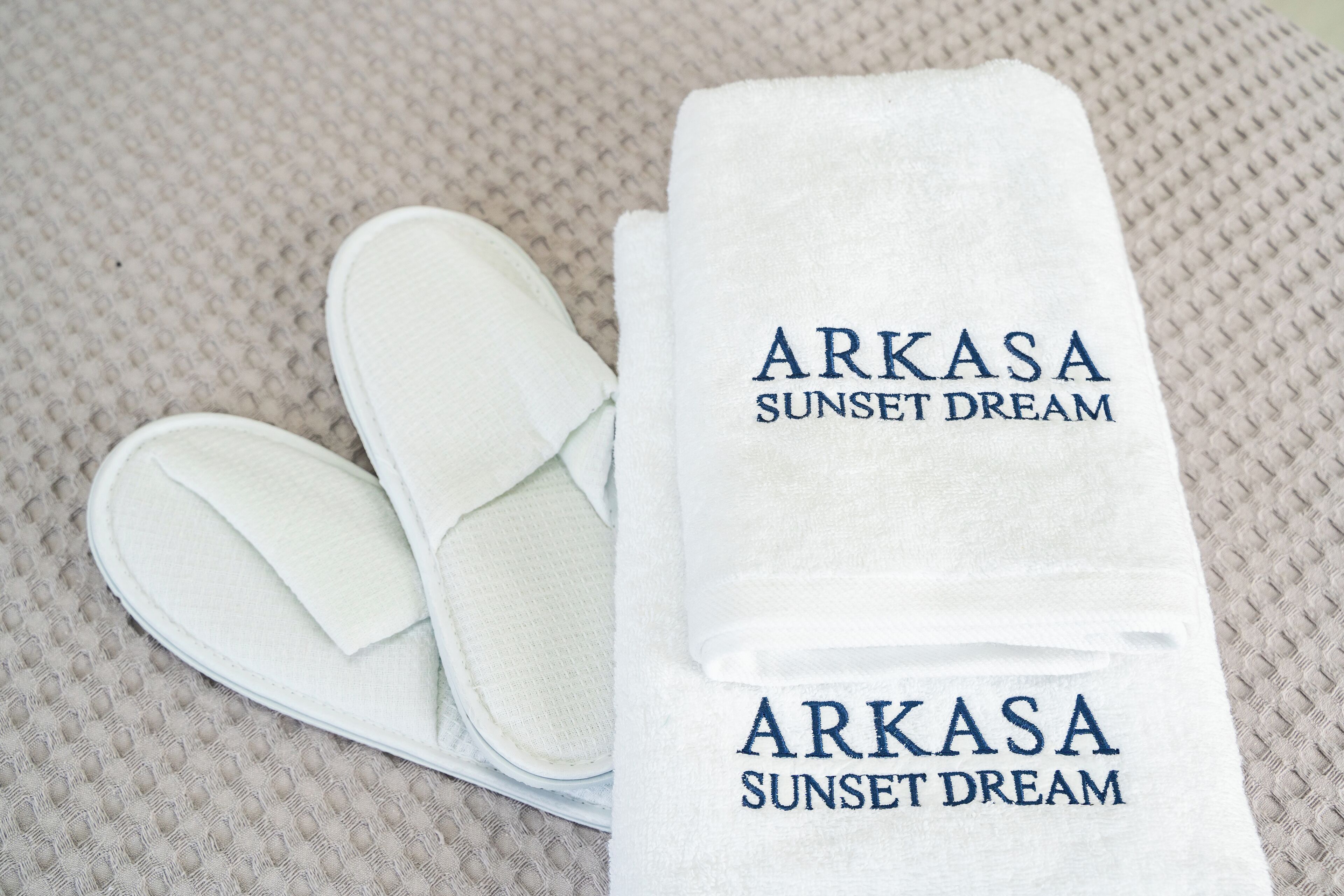 A pair of white slippers and a folded towel with the text "ARKASA SUNSET DREAM" on a textured surface.
