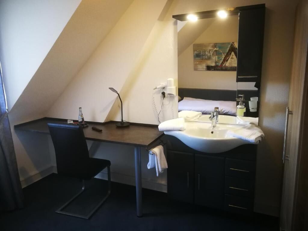 A small, modern bathroom with a dark countertop, a white sink, and a mirror above it. There is a chair and a towel rack visible.