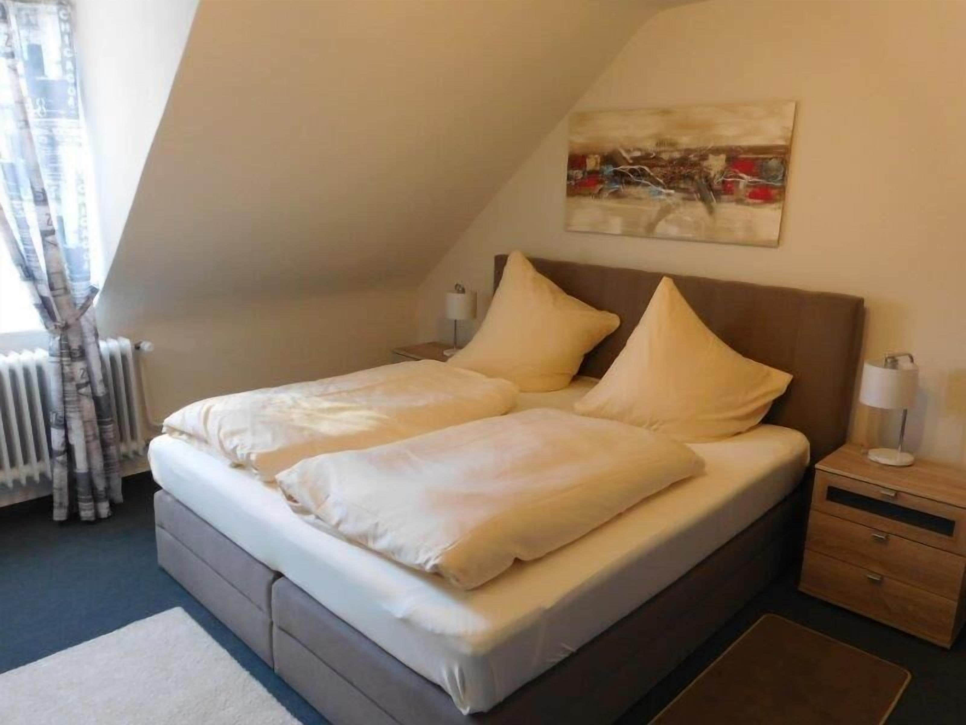 Un dormitorio con cama, mesitas de noche, lámpara, radiador y ventana con cortinas.