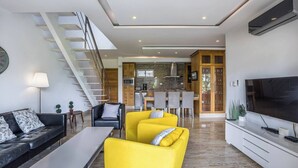 Un salon moderne avec un canapé, des fauteuils jaunes, un téléviseur et un escalier menant à la cuisine.