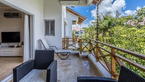 Un balcon doté de meubles en osier, d’un téléviseur et offrant une vue sur un jardin luxuriant.