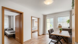 A modern hotel room with a wooden door, a bed with a grey headboard, a small table with a plant, and a view of the outdoors through large windows. - Cozy apartment with 2 bedrooms, private terrace and mountain view (Flattach)
