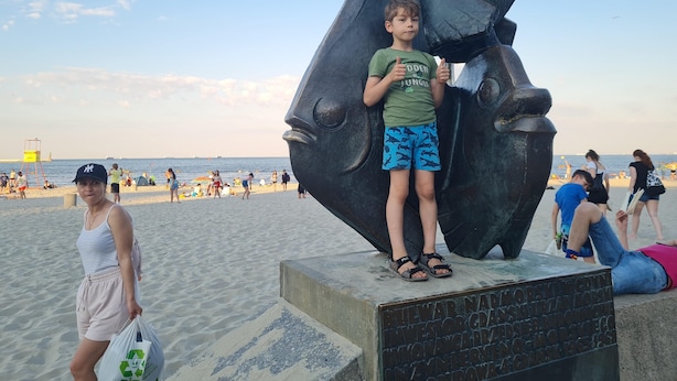 En strandscen med en stor skulptur som föreställer ett fiskhuvud, ett badvakts torn och människor som njuter av stranden.