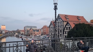En strandpromenad längs floden med en historisk byggnad, en bro och båtar på vattnet.