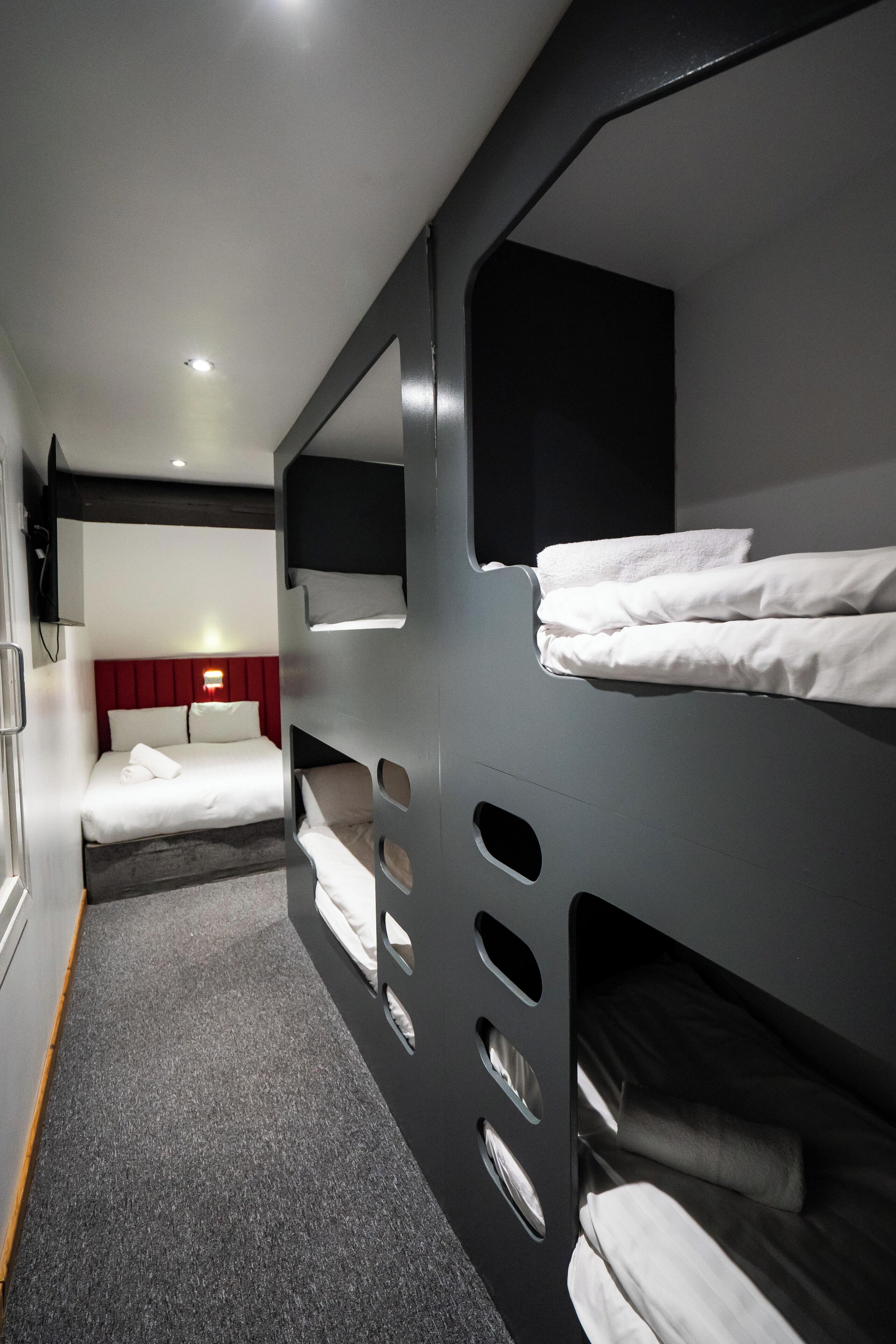 A modern hotel room with bunk beds, a red headboard, a flat-screen TV, and a carpeted floor.