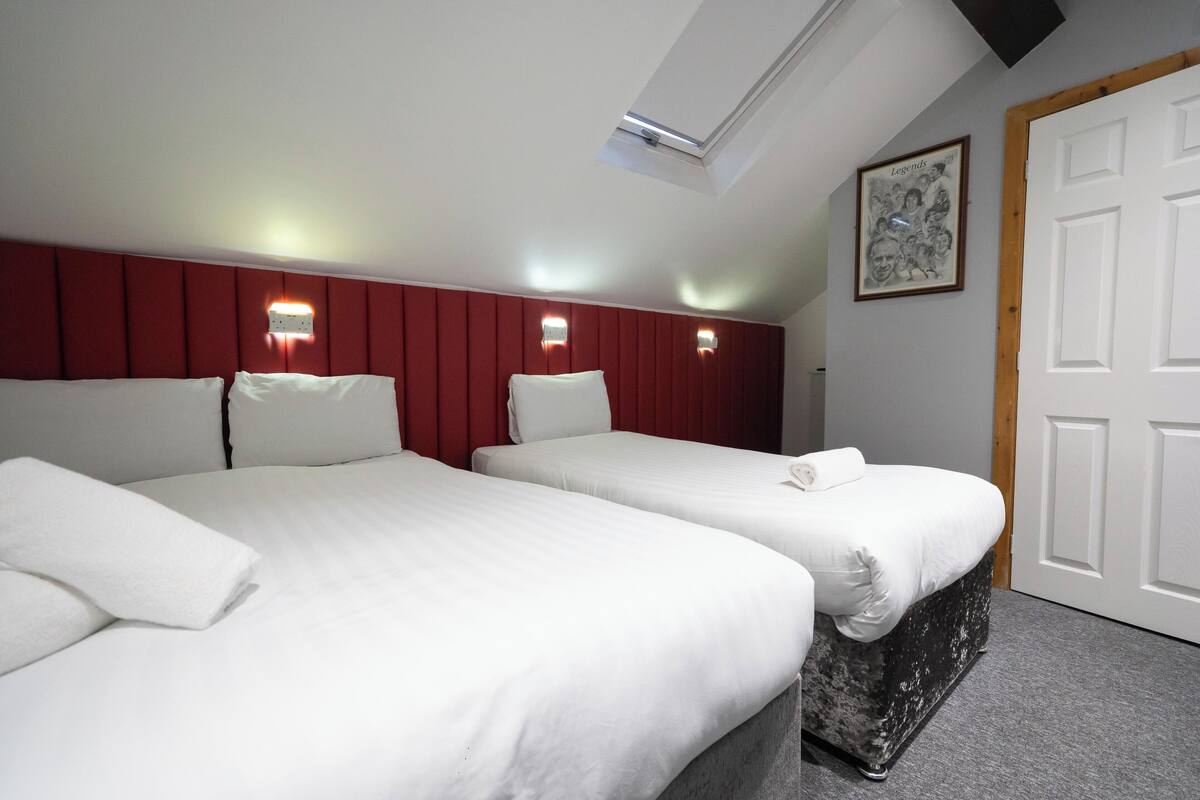 Habitación de hotel con dos camas, cabecera roja, luces fijadas a la pared, tragaluz e imagen en un marco en la pared.