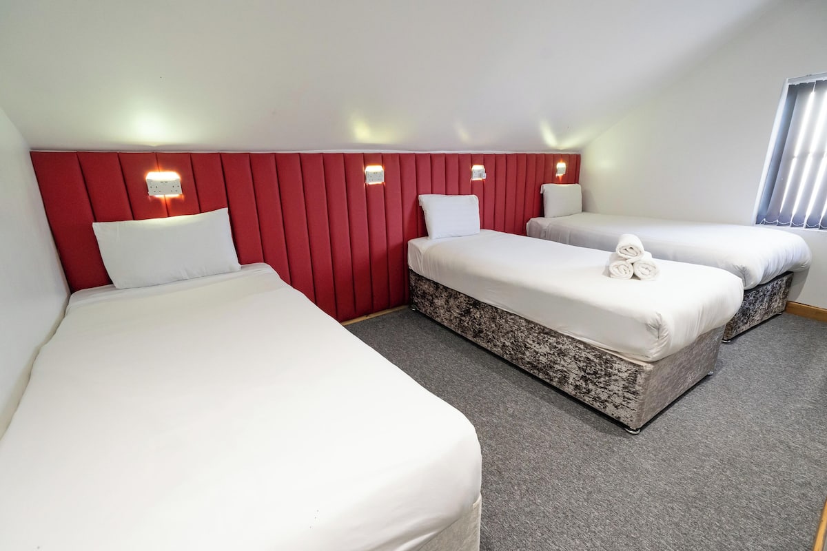 Habitación de hotel con tres camas, un cabecero rojo y dos lámparas fijadas a la pared.