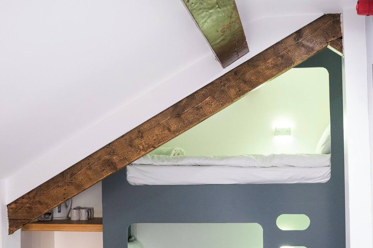 Una habitación tipo dormitorio con literas, techo con vigas de madera y una mesita con un hervidor.