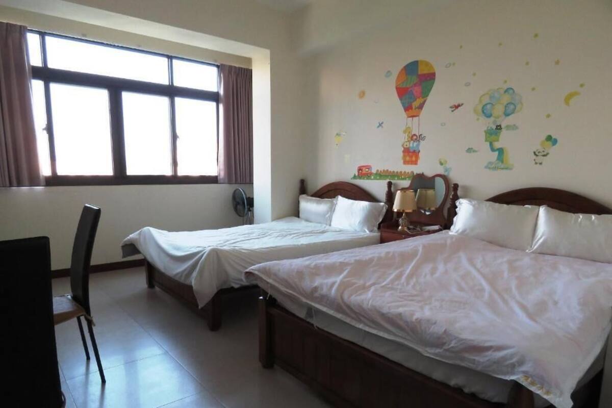 Hotellihuone, jossa on kaksi erillistä sänkyä, tuoli, ikkuna verhoilla sekä seinä, joka on koristeltu lasten tekemillä taideteoksilla.