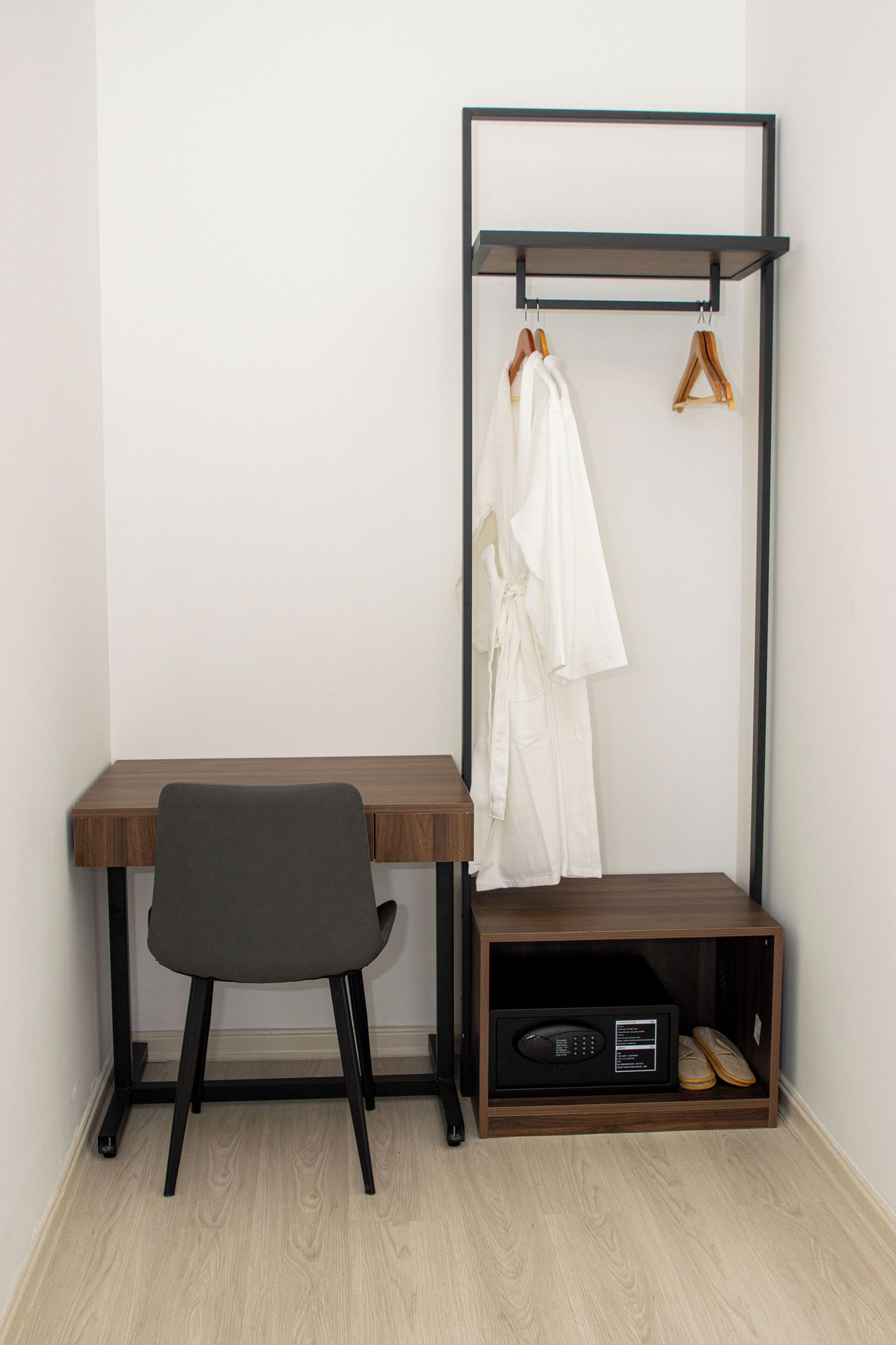 A room with a wooden desk, a chair, a black metal shelf with a white robe and wooden hanger, and a shelf with a radio and shoes.