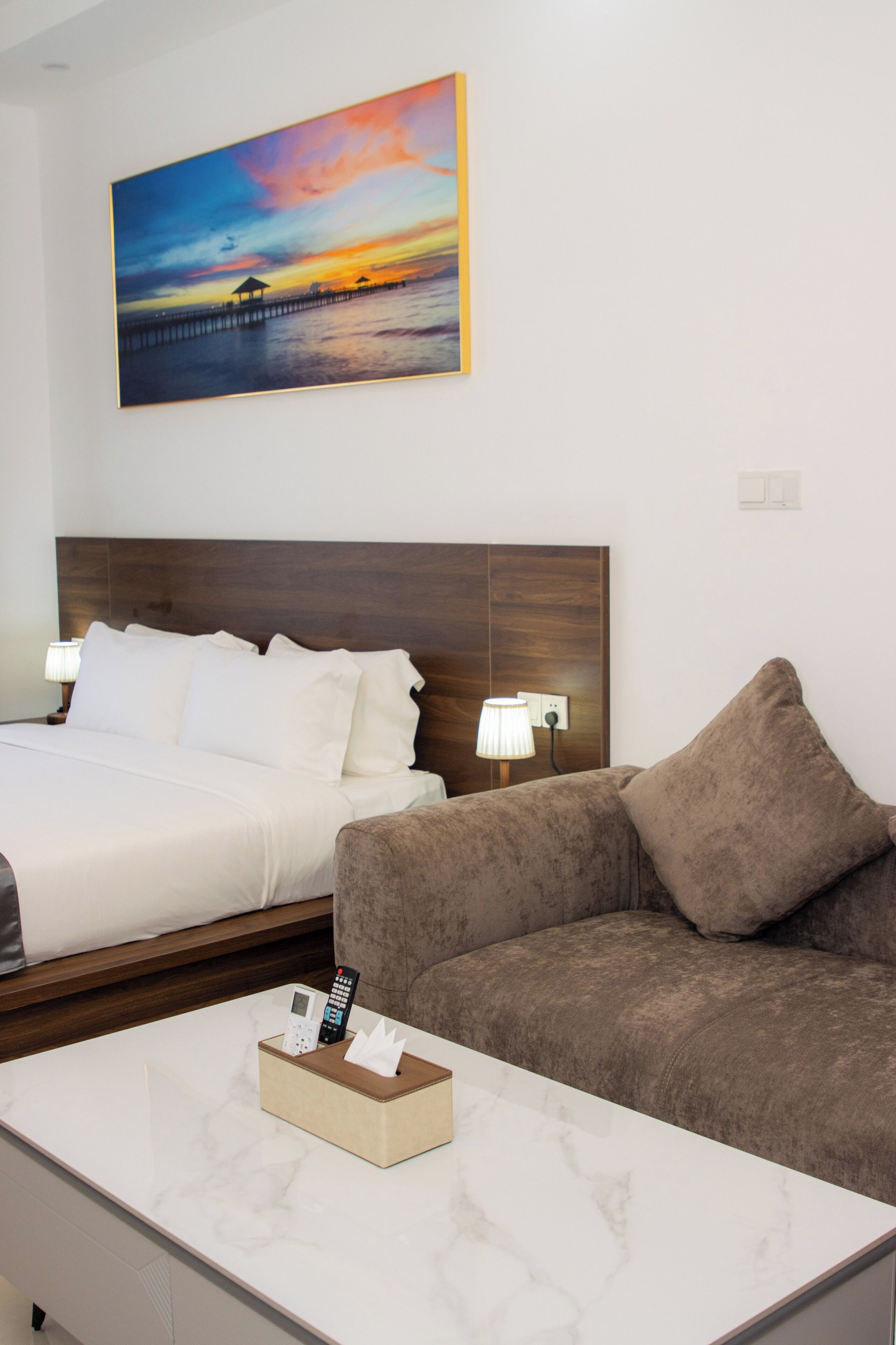 Ein Hotelzimmer mit einem Bett, einer Couch, einem Sonnenuntergangsgemälde und einem Nachttisch mit Lampe und Taschentuchbox.