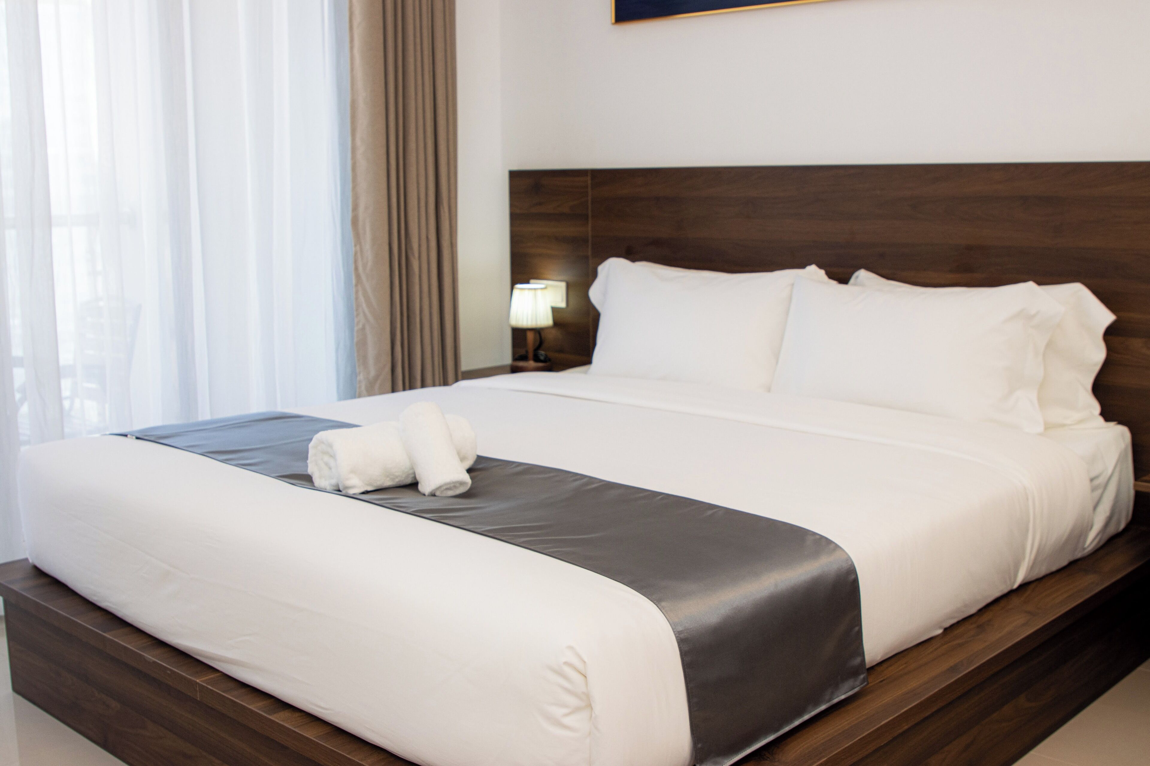 A neatly made bed with white linens and a grey accent, a wooden headboard, and a bedside lamp.