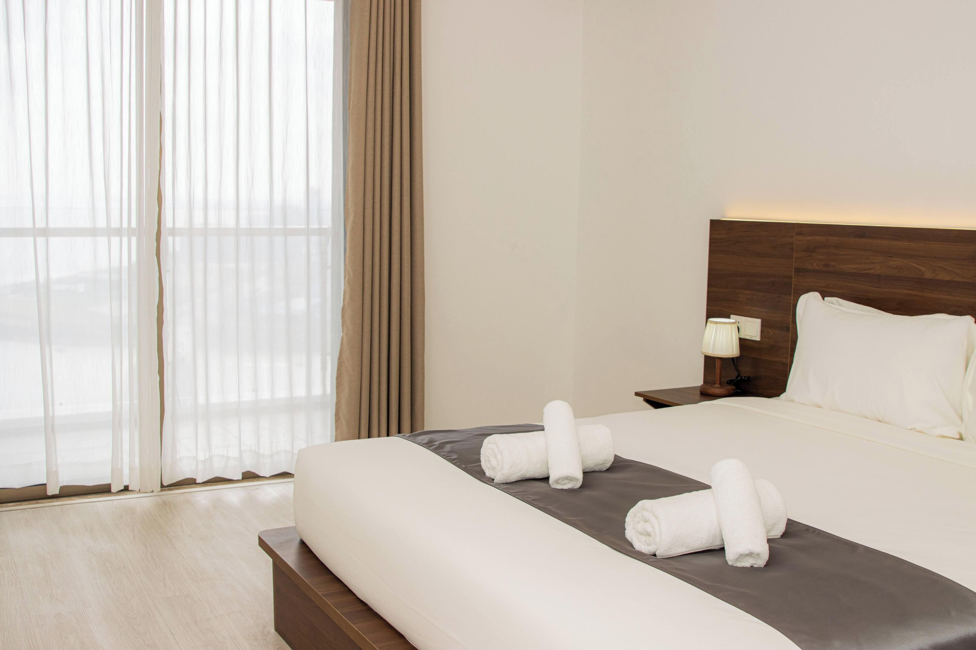 A hotel room with a large bed, a nightstand, a lamp, and a view through sheer curtains.