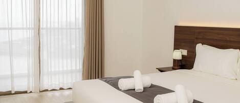 A hotel room with a large bed, a nightstand, a lamp, and a view through sheer curtains.