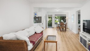 Un salon moderne comprenant un canapé, une table basse en verre, un coin repas avec des chaises, un téléviseur et un coin cuisine avec des étagères et une porte.