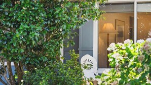 Une fenêtre avec une lampe à l’intérieur, entourée de verdure et d’une chaise blanche.