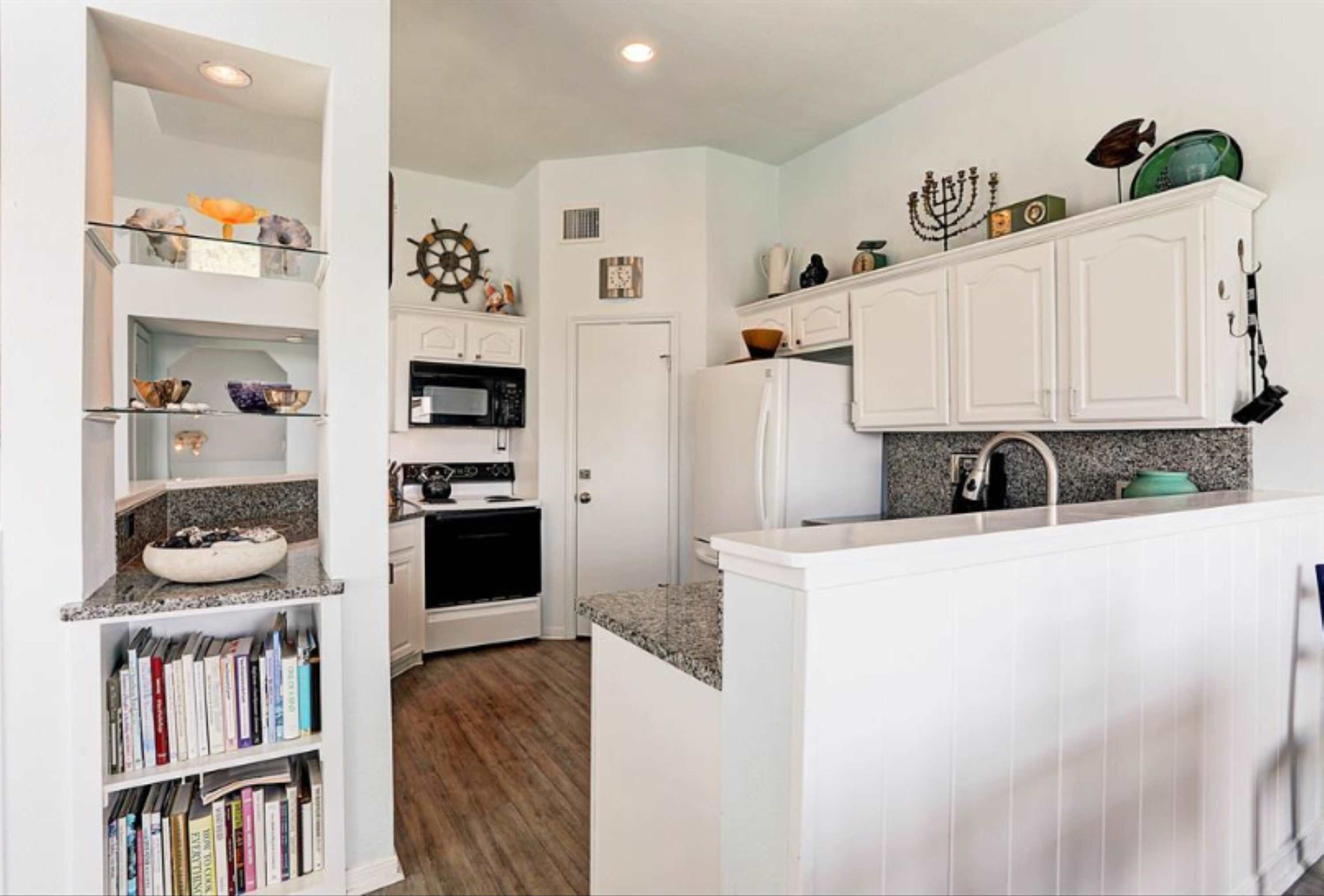 Una cocina con muebles blancos, un microondas, estufa y lavamanos. Hay objetos decorativos en estantes y una estantería.