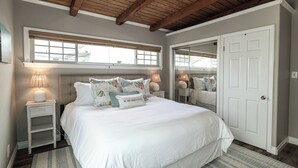 Een slaapkamer met een groot bed, nachtlampjes, een raam met zonwering, een deur en een plafond met zichtbare balken.