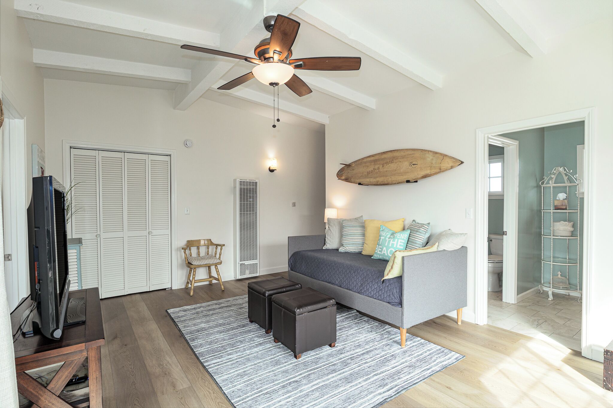 A spacious living room with a ceiling fan, a large bed, a TV, and a wooden surfboard on the wall.