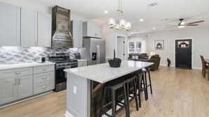 A modern kitchen with grey cabinets, a central island with stools, a built-in oven, and a large window. - The Bella - Modern Equestrian Home with Park Views near Weatherford Square! (Weatherford)