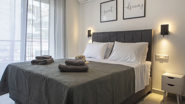 A modern bedroom with a large bed, a bedside table, a wall-mounted lamp, and a window with sheer curtains. - Black & White 2BR Apt steps from Mavili Square (Athina)