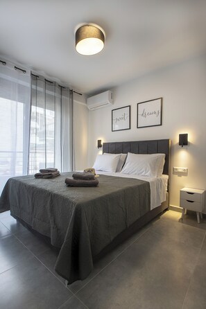 Une chambre à coucher moderne avec un grand lit, une table de chevet, une lampe fixée au mur et une fenêtre ornée de rideaux légers.