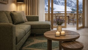 A cozy living room with a green sofa, a round wooden table with candles, and a view of snow-capped mountains through large windows.