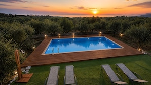 Et rektangulært swimmingpool med træterrasse, omgivet af grønt og med udsigt til solnedgang.