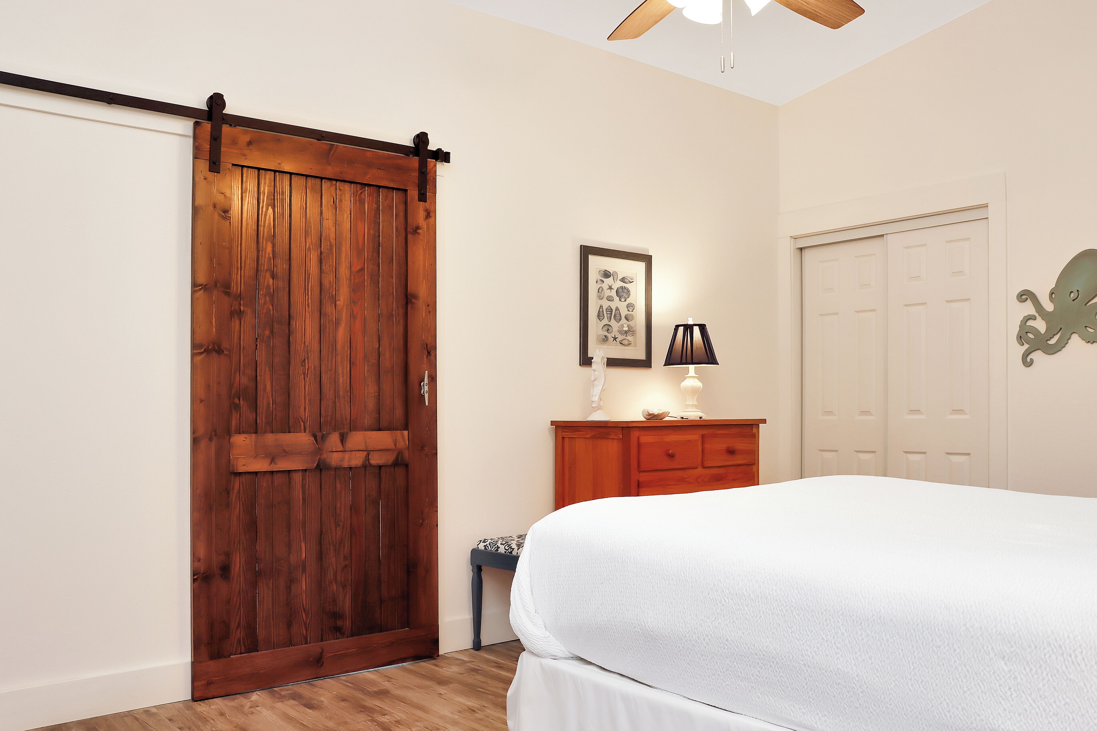 A bedroom with a wooden sliding door, a bed with a white duvet, a bedside table with a lamp, and a wall-mounted picture.