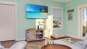 A living room with a television displaying a beach scene, a white door, a small cabinet with board games, and a window with blinds. - ☀️ Short Walk to Beach - Easy Beach Access - Blue Pelican ☀️ (Mexico Beach)