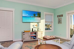 Ein Wohnzimmer mit Fernseher, der eine Strandansicht zeigt, einer weißen Tür, einer kleinen Spielesammlung und einem Fenster mit Jalousien.