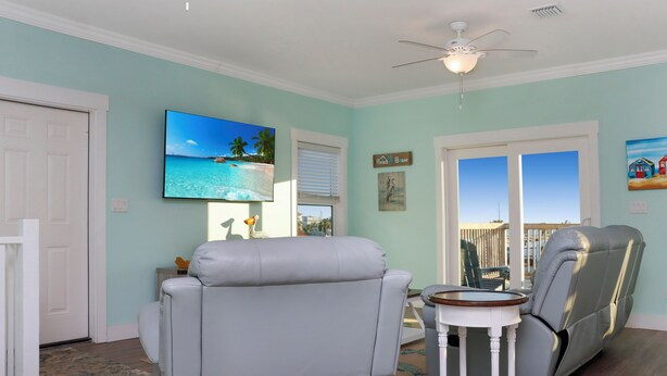 A living room with a gray recliner, a small round table, a ceiling fan, and a large TV on the wall. - ☀️ Short Walk to Beach - Easy Beach Access - Blue Pelican ☀️ (Mexico Beach)