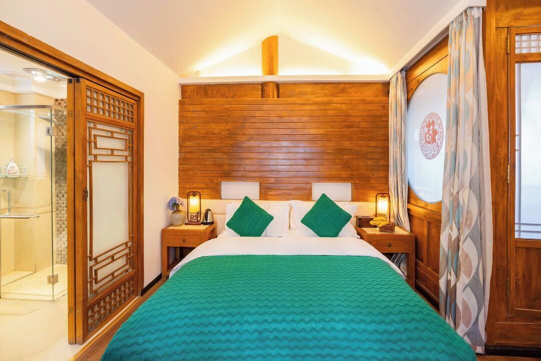Una habitación de hotel con cabecera de madera, una cama con ropa de cama verde, dos mesitas de noche con lámparas y un ventanal con cortinas.