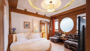 Une chambre dotée d’un grand lit, de poutres en bois, d’un miroir rond, d’une télévision et d’un coin salon avec des meubles recouverts d’un tissu à motifs floraux.