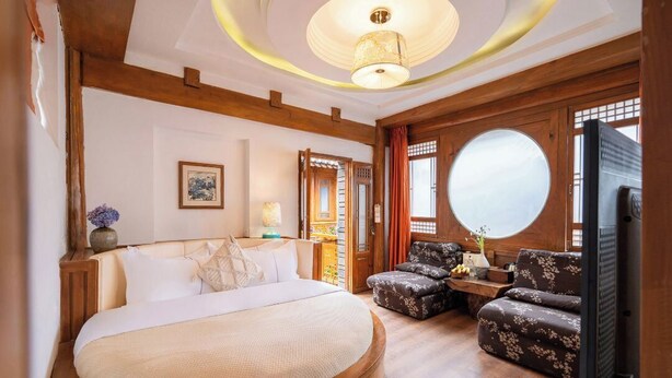 A bedroom with a large bed, wooden beams, a round mirror, a television, and a seating area with floral upholstery. - Shile · A Poetic Guesthouse with Traditional Chinese Charm (Lijiang)