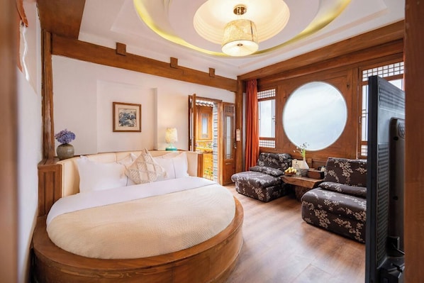 A bedroom with a large bed, wooden beams, a round mirror, a television, and a seating area with floral upholstery. - Shile · A Poetic Guesthouse with Traditional Chinese Charm (Lijiang)