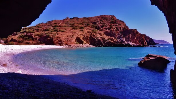 Un paisaje costero visto a través de la entrada de una cueva, que muestra una playa, aguas azules cristalinas y una colina rocosa.