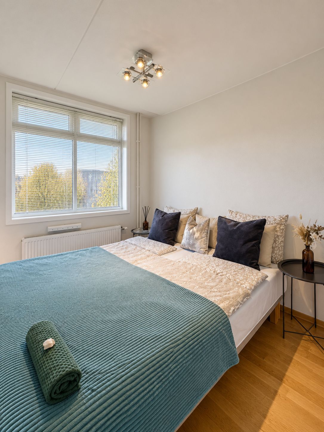 A bedroom with a large bed, a window with blinds, a bedside table with a vase, and a ceiling light fixture.