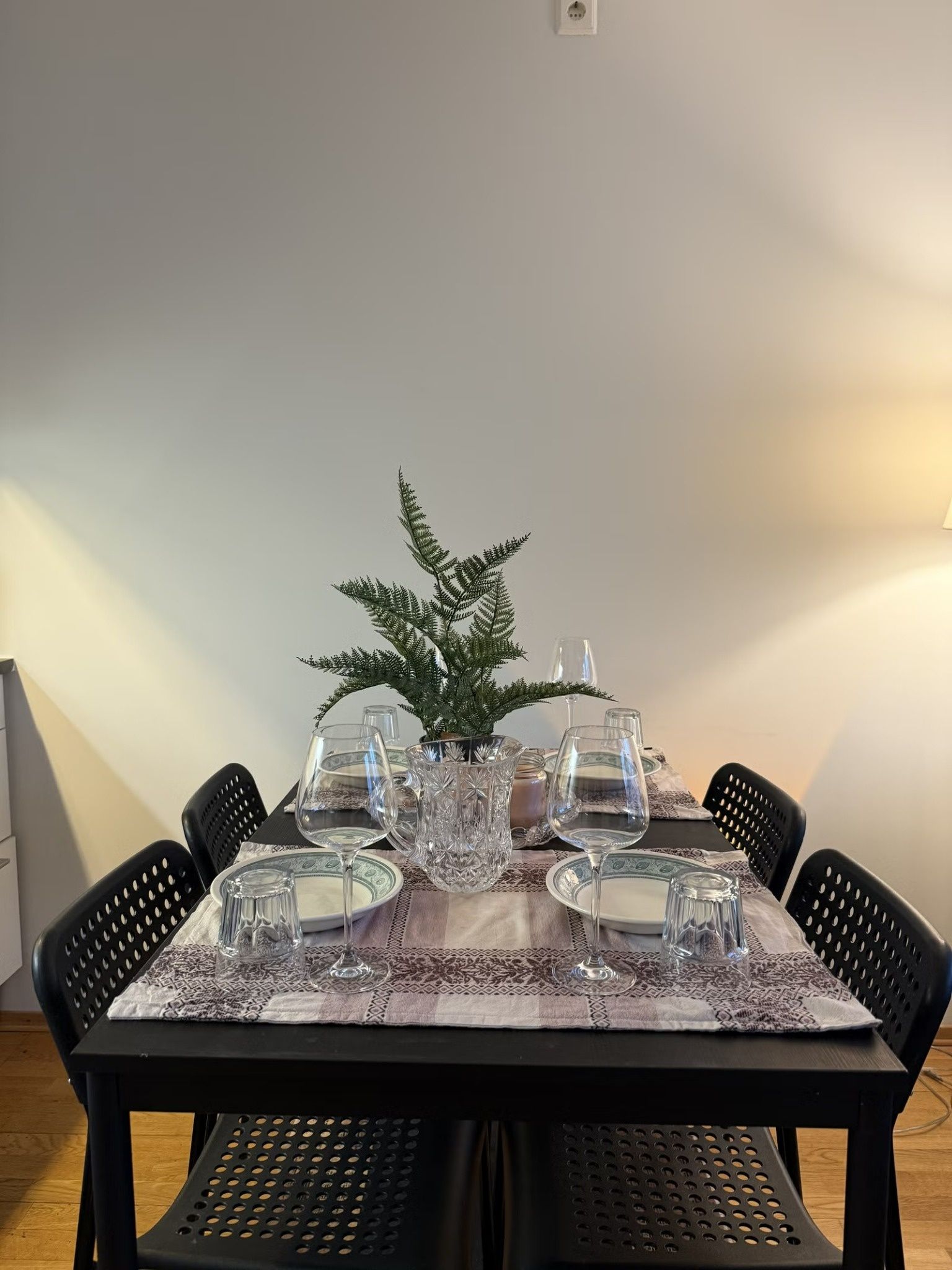 A dining table set for four with clear glassware, a decorative fern, and patterned tablecloth.